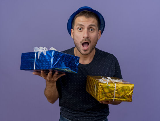 帽子兴奋英俊的白人男子戴着蓝色帽子拿着礼品盒孤立的紫色背景与复制空间持有蓝色男人