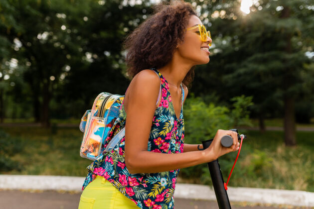摩托车年轻时尚的黑人女子在公园里玩得很开心骑着电动脚踏车在夏天的时尚风格 五颜六色的时髦服装 穿着背包和黄色太阳镜乘坐多彩兴奋