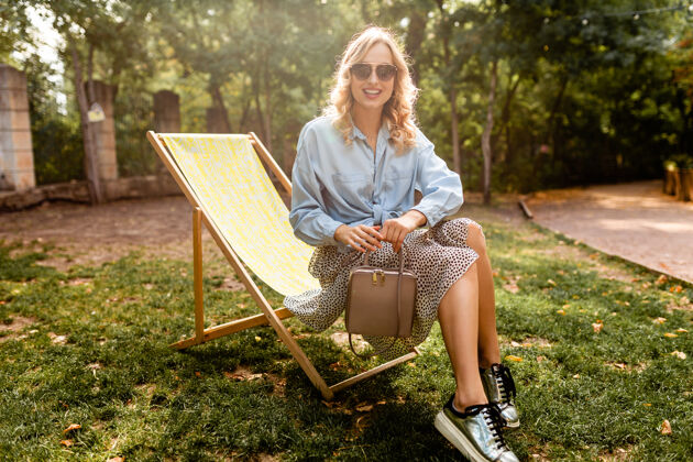 运动鞋迷人的金发快乐的女人坐在躺椅上放松 夏天穿蓝色衬衫 穿着银色运动鞋 优雅的太阳镜和钱包时尚年轻腿