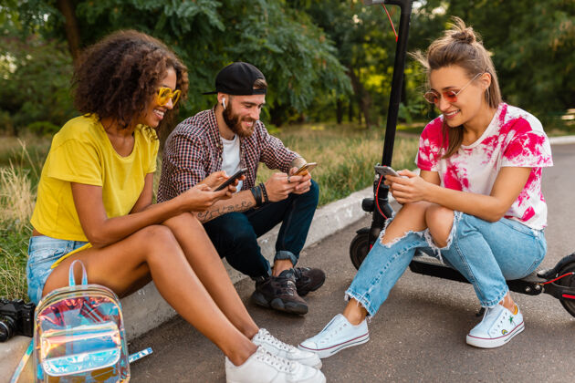 欢呼快乐的年轻朋友们坐在公园里用智能手机微笑 男人和女人一起玩微笑兴奋积极