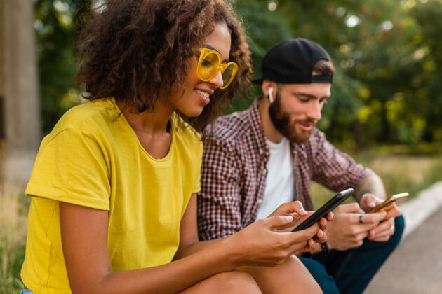 女人快乐的年轻微笑的朋友坐在公园里用智能手机 男人和女人一起玩眼镜团体音乐