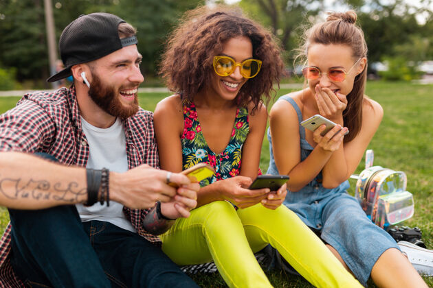 眼镜快乐的年轻朋友们坐在公园里用智能手机微笑 男人和女人一起玩耳机兴奋时尚