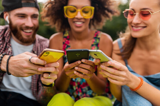 微笑快乐的年轻朋友们坐在公园里用智能手机微笑 男人和女人一起玩情绪无线享受
