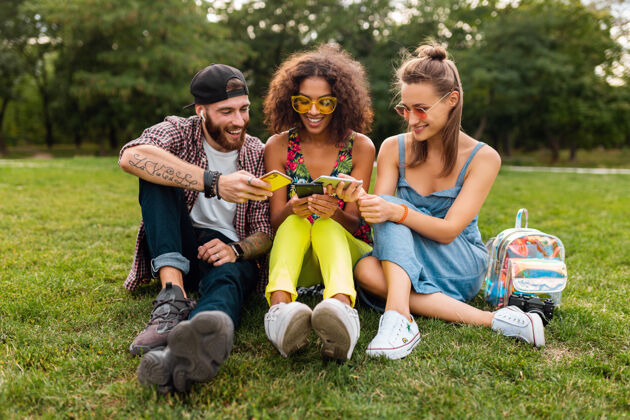 多彩快乐的年轻朋友们坐在公园里用智能手机微笑 男人和女人一起玩手机积极太阳镜