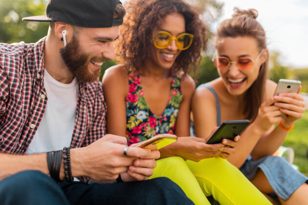 人快乐的年轻朋友们坐在公园里用智能手机微笑 男人和女人一起玩眼镜团体黑色