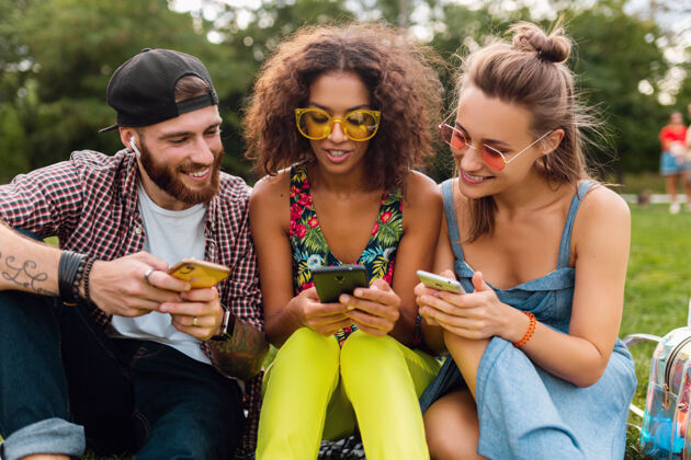 音乐快乐的年轻朋友们坐在公园里用智能手机微笑 男人和女人一起玩多彩手机女人