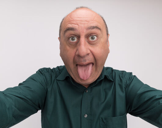 舌头一个快乐的中年男子穿着绿色t恤 拿着相机 在白色的墙上展示着孤立的舌头秀穿T恤