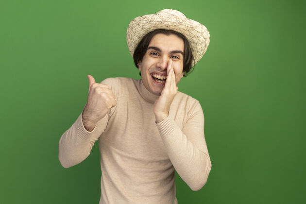 周围面带微笑的年轻帅哥戴着帽子点在一边把手放在嘴边 隔离在绿色的墙上 留着复制空间年轻家伙壁板