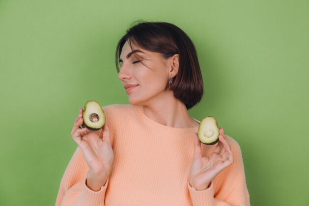异国情调年轻女子穿着休闲桃毛衣隔离在绿橄榄墙上捧着鳄梨 健康护肤理念 复制空间女人饮食模特