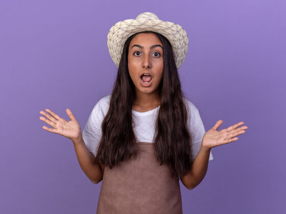 困惑的穿着围裙 戴着夏帽的年轻园丁女孩困惑不解地举起双臂站在紫色的墙上帽子升起年轻的