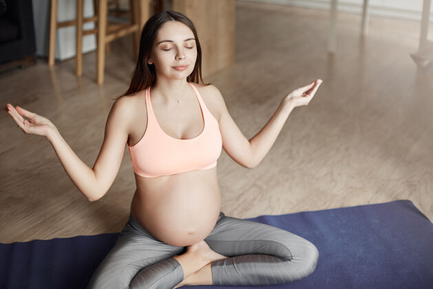 孩子感觉与宝宝和地球的统一冷静迷人的孕妇穿着可爱的运动内衣和紧身裤 坐在瑜伽姿势的滚动垫上 举起手臂在禅宗的姿态 微笑着闭上眼睛健康医疗房子