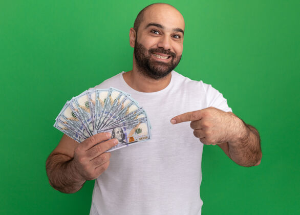 积极一个留着胡须 穿着白色t恤的男人 手里拿着现金 开心地笑着 用食指指着绿墙上的钞票指向现金站