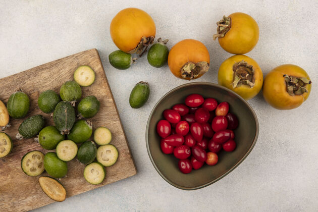 碗整个和一半成熟的feijoas的顶视图被隔离在一个木制的厨房板上 柿子和山茱萸樱桃放在一个灰色表面的碗上可口营养成熟