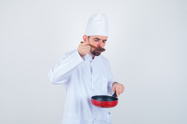 年轻的男厨师身着白色制服 用木勺品尝美食 神情好奇 俯瞰前方家务餐馆站