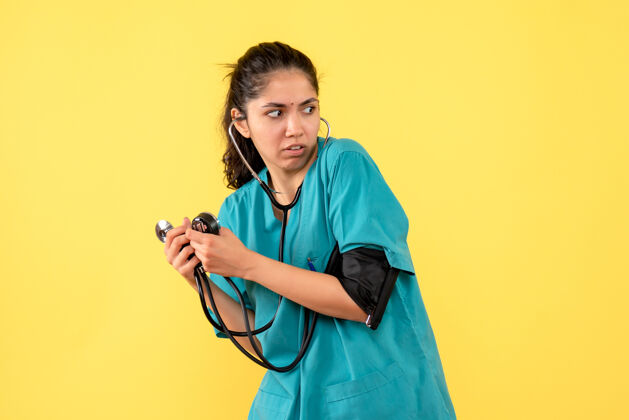 手持前视图吓坏了穿着制服的女医生 拿着血压计站在黄色背景上制服女医生听诊器