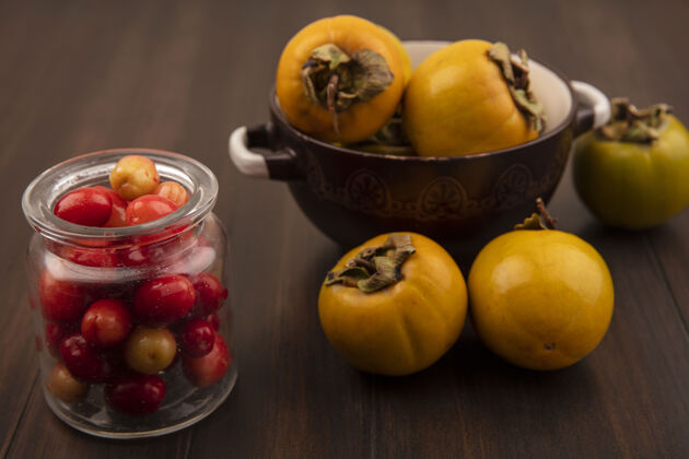 饮食顶视图：红色的科尼莉亚樱桃放在玻璃罐上 柿子水果放在木制表面的碗上配料罐子美味