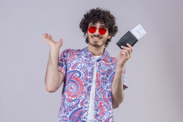 抱着微笑着的年轻英俊的卷发旅行者戴着太阳镜拿着钱包和机票 在孤零零的白墙上空手展示手机票卷发
