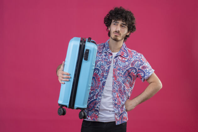 男人未出租的年轻英俊的卷发旅行者男子手持手提箱 手放在腰上 孤立的粉红色墙壁上 有复印空间抱着腰手