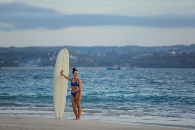 人美丽的女孩拿着冲浪板站在沙滩上太阳女人海洋
