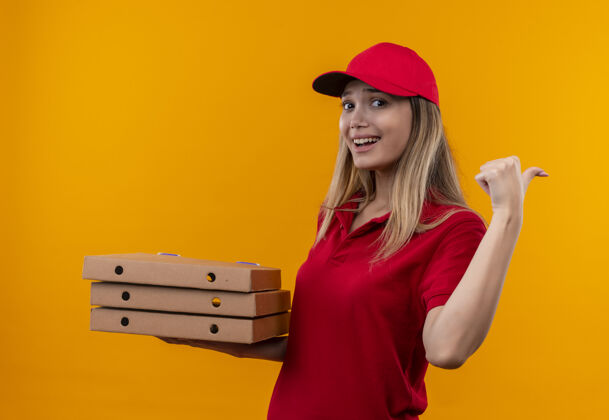 壁板面带微笑的年轻送货女孩身穿红色制服 戴着帽子 手里拿着披萨盒 指着隔离在橙色墙上的一侧抱送货穿