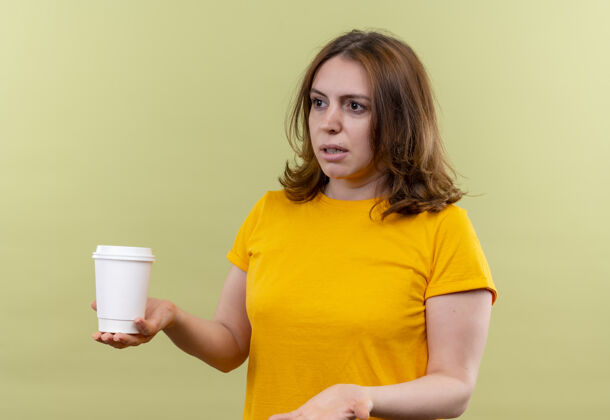 炫耀的年轻随意的女人拿着塑料咖啡杯 在隔离的绿色墙壁上空手展示手杯子咖啡