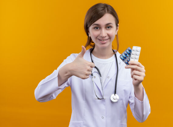 女人面带微笑的年轻女医生穿着医用长袍 手持医用药物的听诊器 在孤立的橙色墙上竖起大拇指长袍年轻人微笑
