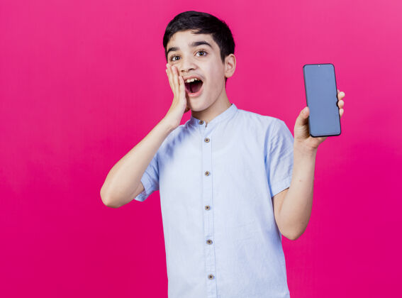 男孩惊讶的年轻白人男孩展示手机 看着相机 手放在脸上 在深红色的背景下 与复制空间隔离脸惊喜手