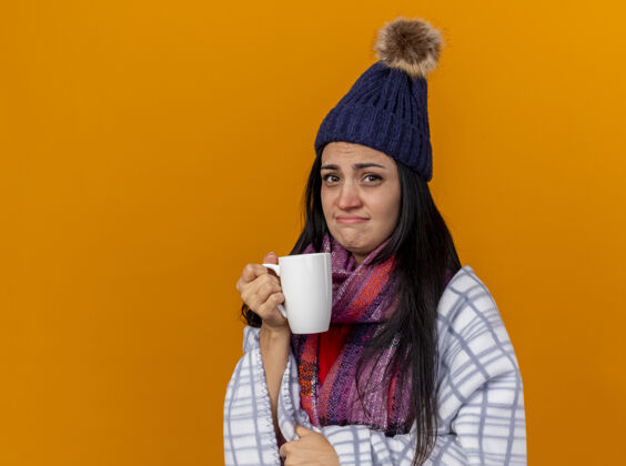 疾病虚弱的年轻白种人生病的女孩戴着冬天的帽子 围巾裹着格子布 手里拿着一杯茶 看着隔离在橙色背景上的相机 还有复制空间帽子茶年轻