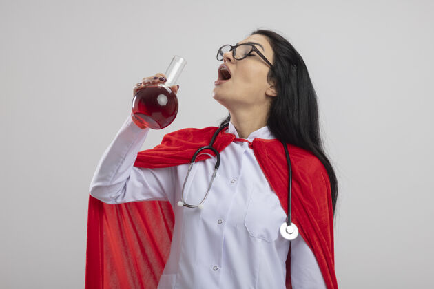 听诊器年轻的白种人超级英雄女孩戴着眼镜和听诊器拿着装有红色液体的化学瓶准备喝它 闭着眼睛隔离在白色背景上 留有复印空间持有液体年轻