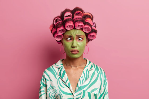 风格搞笑的民族女青年做鬼脸 交叉眼睛 涂发卷 为特殊的日子做发型 脸上戴绿色保湿面膜民族绿色面具
