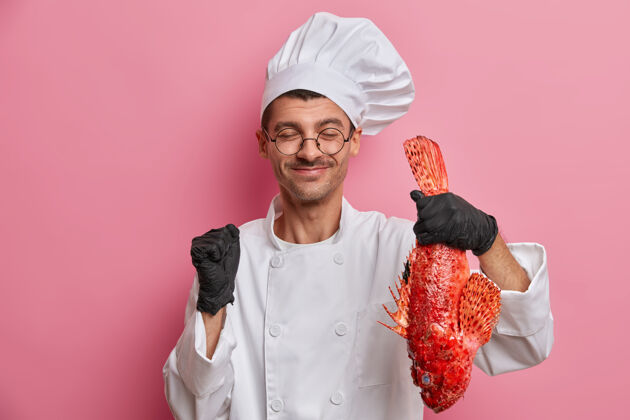 拳头准备海鲜穿着厨师制服的快乐的欧洲厨师 橡胶手套抱着红海鲈鱼 高兴地握紧拳头专业快乐制服