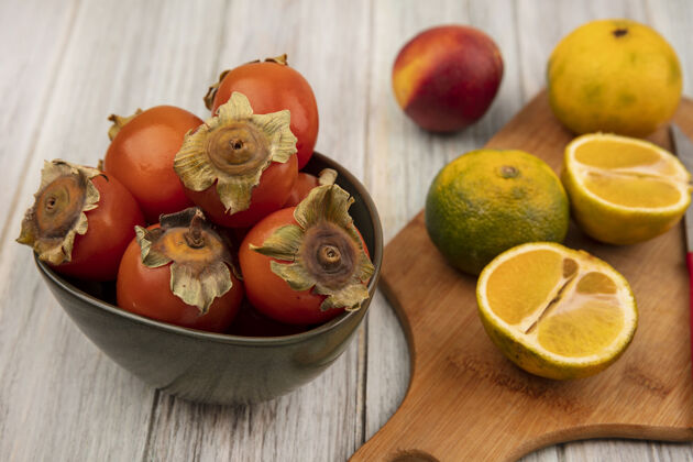 新鲜木制菜板上橘子的俯视图 柿子放在碗上 桃子被隔离在灰色的木墙上木板食物视野