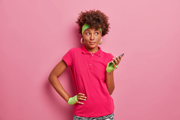 人物运动型的深色皮肤卷发女士手放在腰上 手拿手机 使用在线训练专用应用程序 戴着头带 运动手套 粉色t恤运动员健身室内