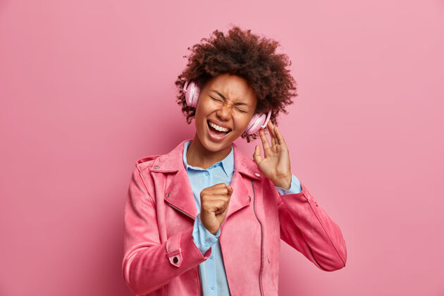女性快乐的美国黑人妇女手牵麦克风 高声唱歌 头戴耳机听音乐 歪着头 傻乎乎的 穿着时髦的夹克粉色音频女士