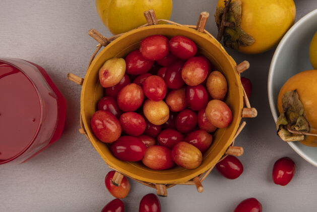 椭圆形浅红色和椭圆形的山茱萸水果在桶上的顶视图 柿子水果在碗上 新鲜的山茱萸果汁在灰色的墙上的玻璃杯里柿子视图苍白