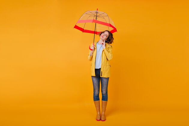 伞红色阳伞下站在黄色墙壁上沉思的浪漫女孩的全长肖像穿着牛仔裤和秋鞋的时尚女模特一边打着伞一边看着远处的摄影棚镜头黑发季节室内