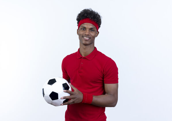 男带着微笑的年轻的非裔美国人运动男子戴着头带和手环拿着球孤立在白色运动腕带白