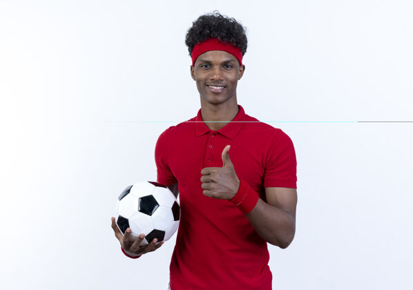 男微笑着的年轻的美国黑人运动男子戴着头带和腕带拿着球他的大拇指孤立在白色球年轻白