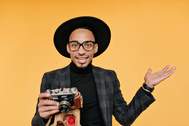 时尚一个戴着黑帽子的惊讶男摄影师一张非洲年轻人的室内照片 相机被隔离在黄色的墙上情感男孩装备