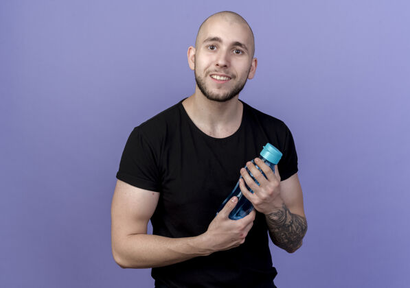 瓶子带着微笑的年轻运动男子拿着水瓶孤立地站在紫色地上年轻水运动