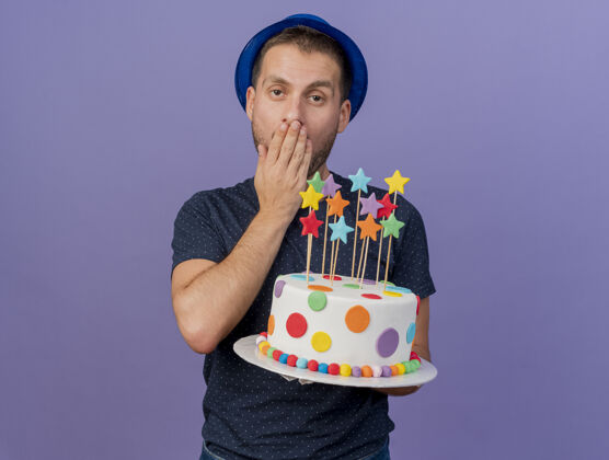 人戴蓝帽子的帅哥生气地把手放在嘴上 把生日蛋糕孤立地放在紫色的墙上蛋糕穿着手