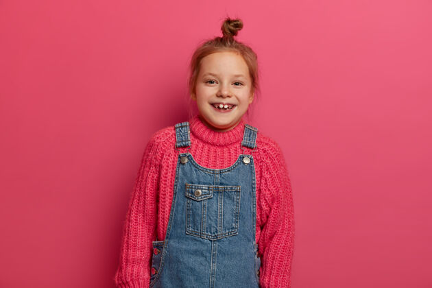 乐趣满脸笑容的小宝宝 发髻 穿着针织毛衣和牛仔布 看起来很高兴 在粉红色的墙上摆姿势 要和孩子们一起玩情绪 孩子们童年脸模特