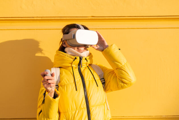 玩家一个戴着虚拟现实眼镜的女人的肖像 在冬天的阳光下 穿着暖和的衣服 靠在黄色的墙上模拟微笑Vr眼镜