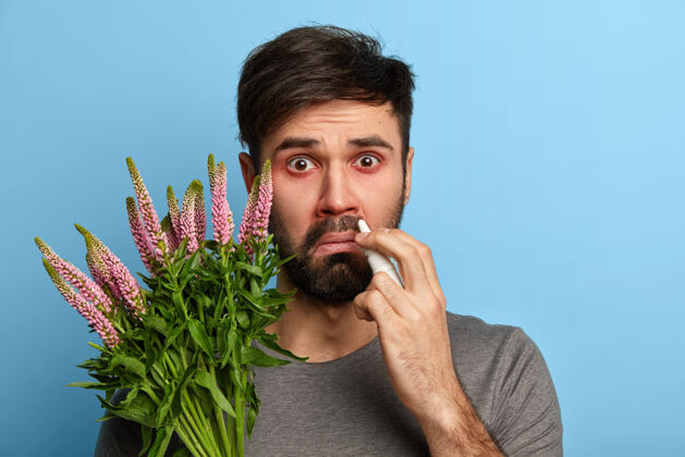 不高兴胡须不健康的男人患有季节性过敏 喷鼻涕 抱着植物 对过敏原敏感 对着蓝色墙壁摆姿势医学概念年轻疼痛痛苦