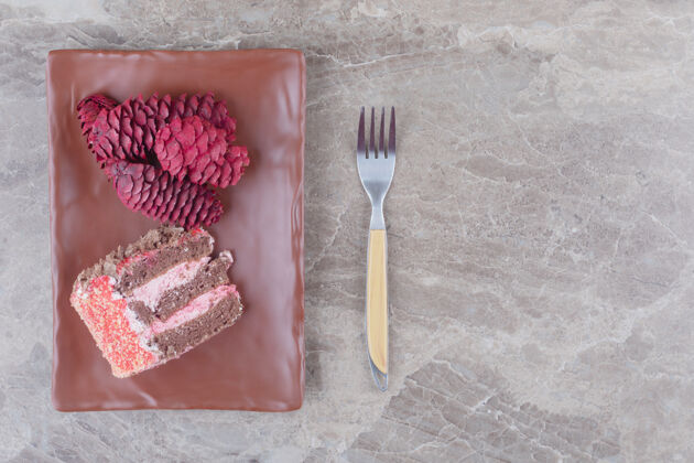 蛋糕一块蛋糕和红松球果放在一个盘子里 旁边是一个大理石叉子蛋卷美味松树