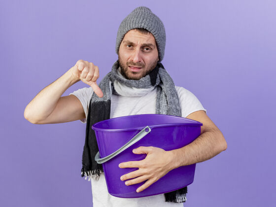 疾病一个戴着冬天帽子 戴着围巾 拿着塑料筐 拇指朝下的年轻人 被隔离在紫色的背景上帽子不愉快塑料