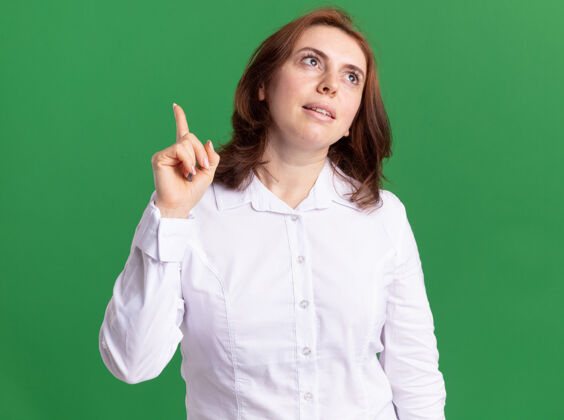 女人身穿白衬衫的年轻女子面带微笑地抬起头来 伸出食指思考着站在绿色的墙上立场年轻脸