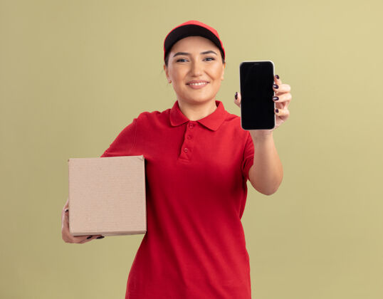 交货年轻的送货员身穿红色制服 戴着帽子 手里拿着一个纸板箱 手里拿着智能手机 站在绿色的墙上 面带微笑地看着前面帽子纸板年轻