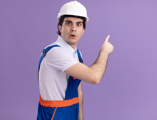 人年轻的建筑工人穿着建筑制服 戴着安全帽 看上去很困惑 用食指指着站在紫色墙上的一边侧线制服立场