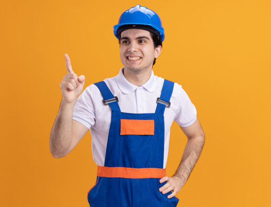 微笑年轻的建筑工人穿着建筑制服 戴着安全帽 一边微笑一边自信地用食指指着站在橙色墙上的东西年轻人建筑什么